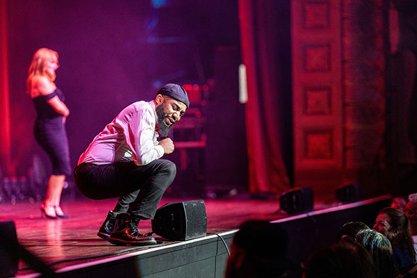 Mott, Motown and More event photo of a man at the front of stage crouching down and interacting with the seated audience; a woman in a black dress appears in the background.